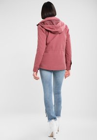 Donna con capelli corti e scuri, che indossa una giacca a vento rosa con cappuccio, jeans azzurri sfilacciati e Stivaletti bianchi con tacco, in piedi di fronte a uno sfondo neutro.