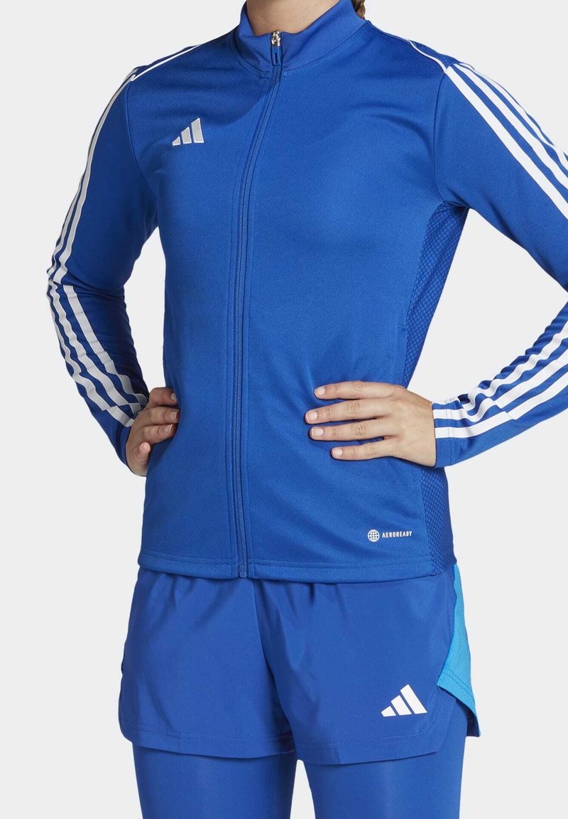 Henkilö pukeutuneena siniseen Adidas-vedettävään takkiin, jossa on valkoiset raidat hihoissa, ja vastaaviin sinisiin shortseihin, joissa on Adidas-logo ja Aeroready-teksti.