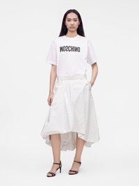 Γυναίκα που φοράει λευκό t-shirt Moschino και ασύμμετρη λευκή φούστα με λεπτές ρίγες, καθώς και μαύρα ψηλοτάκουνα με λουράκια, στέκεται μπροστά σε απλό φόντο.
