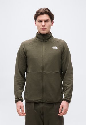 Ung mand vendt fremad iført en olivengrøn North Face jakke med fuld lynlås og matchende bukser mod en ensartet lys baggrund.