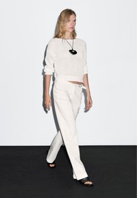 Witte gebreide trui, lichtbeige wijde broek met riemdetails en zwarte open-toe sandalen, gedragen door een model tegen een effen achtergrond.