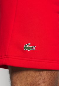 Röda idrottsshorts med en slät textur och en vit kanten grön krokodil-logotyp på fållen. Sytt detaljer vid sömmarna.