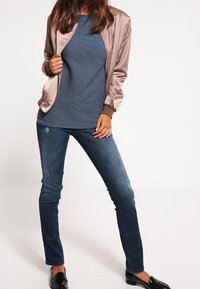 Světle růžová saténová bomber jacket přes námořnický top s ribbed vzorem, spárovaná s úzkými tmavě modrými džíny a černými mokasínami.