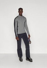 Grijs atletisch shirt met lange mouwen en ritssluiting bij de hals, met contrasterende donkere mouwen; gecombineerd met marineblauwe broek en zwarte skischoenen, ski-uitrusting vasthoudend.