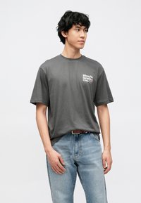 Sivé bavlnené tričko s krátkymi rukávmi, okrúhlym výstrihom a potlačou na prednej strane hrudníka. Kombinované so svetlo modrými rifľovými nohavicami.