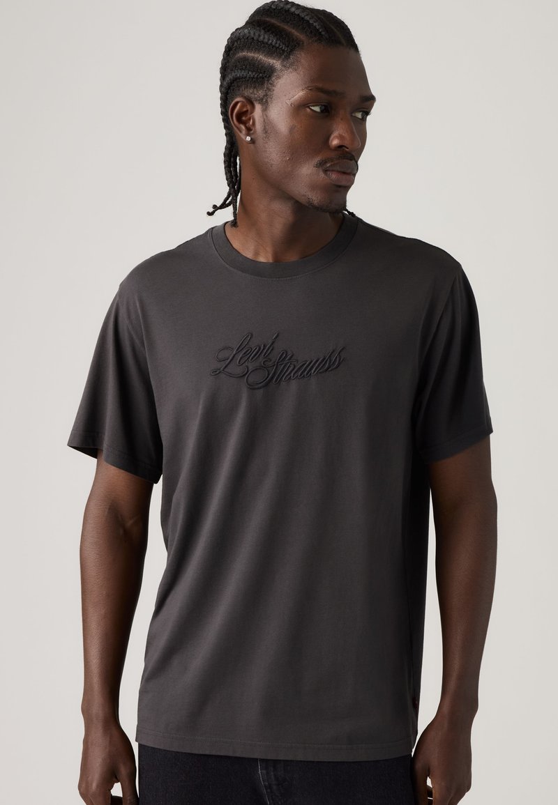 Man with braided hair wearing a dark gray Levi Strauss t-shirt looking to the side against a plain light background.