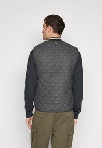 Gray quilted vest with a textured pattern, featuring a yellow logo accent near the collar, worn over a black long-sleeve shirt and green cargo pants.
