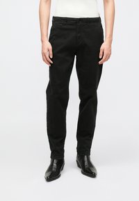 Black cotton chinos with a straight fit, featuring a button closure, side pockets, and a smooth texture. Paired with black cowboy boots.