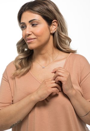 Beige V-Ausschnitt-Shirt mit kurzen Ärmeln, ergänzt durch eine zarte goldene Halskette mit rechteckigem Anhänger und kleinen Steckerohrringen.