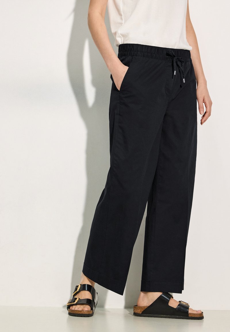 Pantaloni neri a gamba larga con cintura elastica e coulisse, dotati di tasche laterali. Abbinati a sandali neri e a una maglietta di colore chiaro.