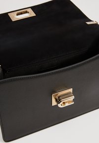 Sort læderhåndtaske med en struktureret finish, der har en guld hardware-lås og en lynlåslomme indeni. Moderne rektangulær form.