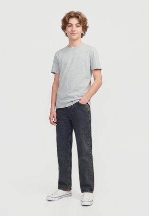 Adolescent debout, portant une chemise à manches courtes gris clair, un jean foncé et des baskets blanches, avec une main dans sa poche.