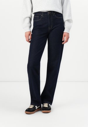 Persoon met donkerblauwe rechte jeans, lichtgrijze trui en zwarte sneakers met witte veters en gumzolen, staand tegen een witte achtergrond.