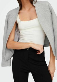 Grå texturerad jacka med en strukturerad design, bärs öppen över en vit tanktop, tillsammans med högmidjade svarta byxor med knappdetalj.