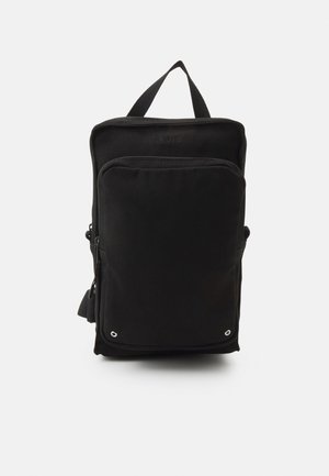 Mochila de lona negra con forma rectangular, que presenta un bolsillo con cremallera en la parte frontal, un asa en la parte superior, una correa para el hombro ajustable y detalles metálicos ventilados.