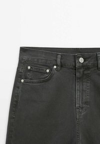 Zone de la taille d'un jean en denim noir montrant le bouton avant, les passants de ceinture et une petite poche avant avec des rivets métalliques.