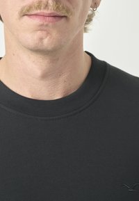 Schwarzes Baumwoll-Sweatshirt mit Rundhalsausschnitt, gerippten Bündchen und kleinem gesticktem Logo auf der unteren rechten Seite. Glatte Textur.