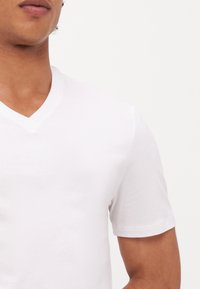 Weißes Baumwoll-T-Shirt mit V-Ausschnitt, kurzen Ärmeln, glatter Textur und gefitteter Form, das einen klaren, minimalistischen Look zeigt.