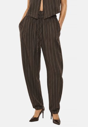 Persona indossa pantaloni marroni a righe larghe con cintura annodata, gilet coordinato, mani nelle tasche e décolleté nere a punta, su uno sfondo neutro.