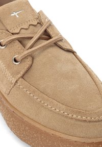 Beige Wildlederschuh mit kontrastierenden gesteppten Akzenten und einer Gummisohle. Verfügt über Metallösen und dekorative Schnitträtsche.