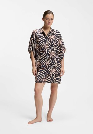 Frau steht barfuß und trägt ein locker sitzendes, knielanges Kleid mit Tiermuster, Kragen und dreiviertellangen Ärmeln vor weißem Hintergrund.