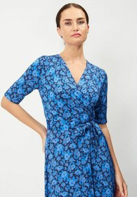 Sininen kukallinen kietaisumekko lyhyillä hihoilla, jossa on syvä V-kaula-aukko ja nauhavyötärö. Kangas on teksturoidulla kuosilla vaihtelevissa sinisen sävyissä.