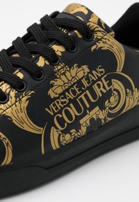 Black sneaker with gold ornate patterns and "VERSACE JEANS COUTURE" text. Features black textured laces and rubber sole.