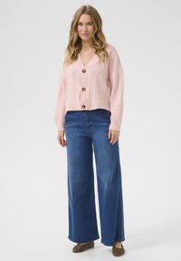 Světle růžový pletený cardigan s výstřihem do V, který má strukturovaný vzor a dřevěné knoflíky, v kombinaci s modrými džínami s širokými nohavicemi.