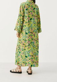 Robe maxi en tissu vert vif arborant un motif floral en rose et bleu. Manches longues, col et silhouette fluide. Portée avec des sandales tan.