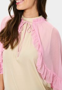 Pink sheer cape with ruffle accents, open front design, paired with beige top featuring drawstring at the neckline. Both fabrics are lightweight.