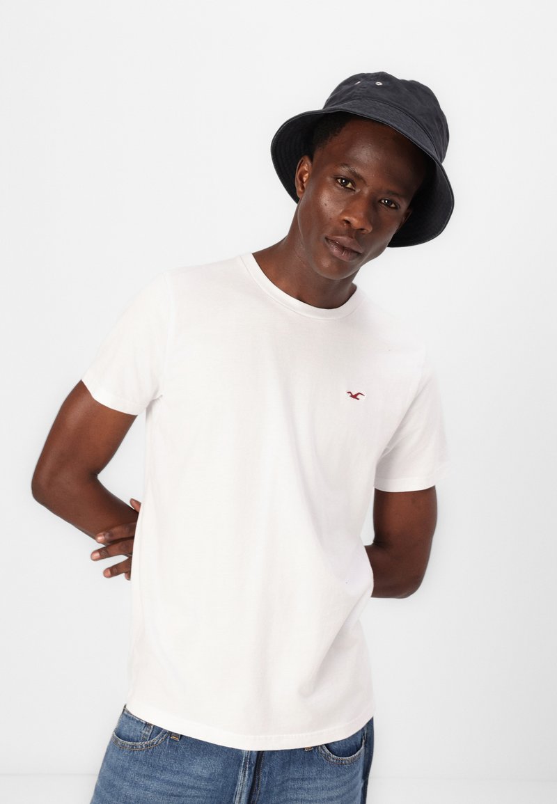 Weißes Baumwoll-T-Shirt mit rundem Halsausschnitt und kurzen Ärmeln. Ein kleines rotes Logo ist auf der linken Brust sichtbar. Kombiniert mit einem dunklen Bucket Hat.