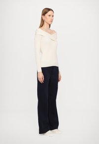 Sweater de malha canelada off-white com ombros à mostra, combinado com calças largas navy e ténis brancos, apresentando um corte descontraído e uma textura suave.