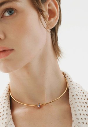 Collar de choker de metal dorado con forma circular lisa y un pequeño acento colorido. Lleva puesto alrededor del cuello, combinado con un cuello blanco texturizado.