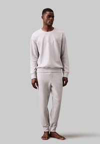 Grijze sweatshirt en bijpassende broek gemaakt van zachte stof, met een ronde hals en geribbelde manchetten. Model staat blootsvoets tegen een egale achtergrond.