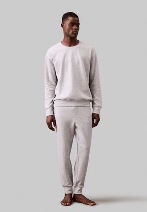 Grau sweatshirt und passende Hose aus weichem Stoff, mit Rundhalsausschnitt und gerippten Bündchen. Das Model steht barfuß vor einem schlichten Hintergrund.