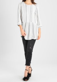 Hvid tunika med sorte lodrette striber; flare-design. Sorte slidte jeans med perleaccenter; parret med sorte strophæle.