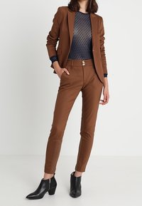 Blazer marrón ajustado con un acabado suave, combinado con una blusa de malla azul marino con puntos y pantalones marrones ajustados. Botines negros completan el conjunto.