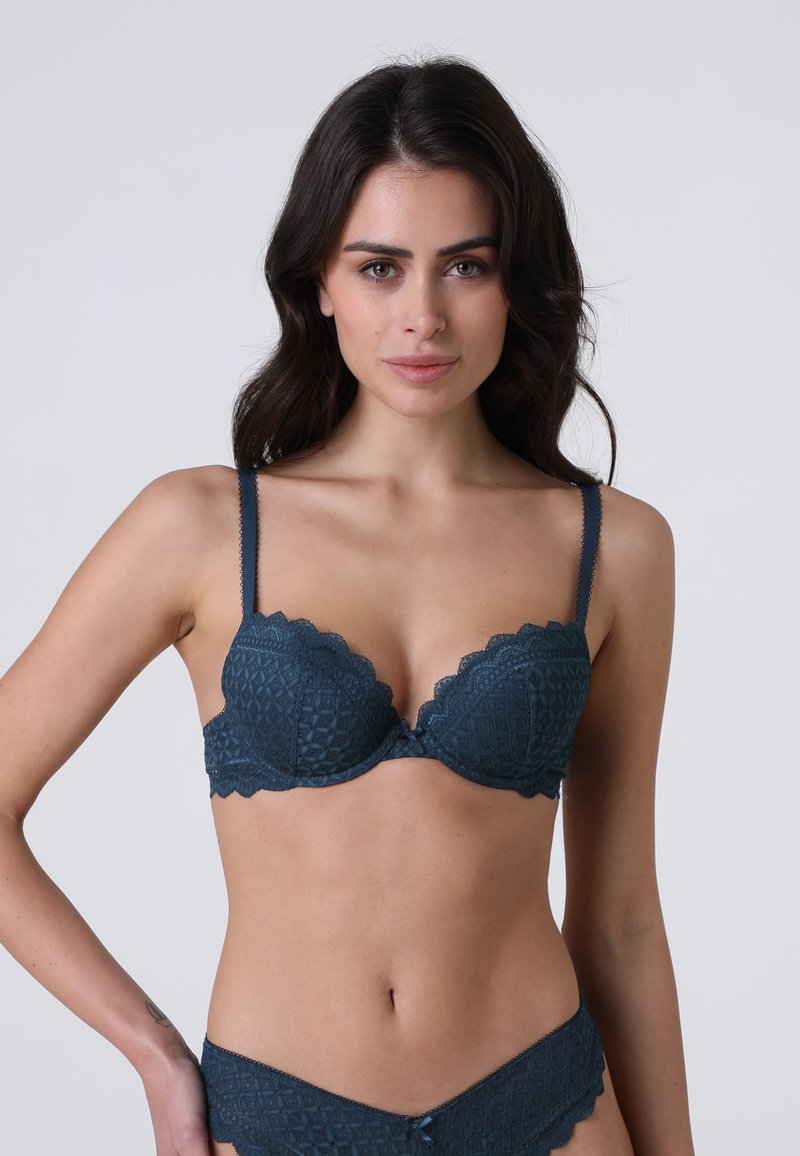 Donna con capelli scuri che indossa un reggiseno e delle mutandine in pizzo di un abbinato color teal scuro, posando con una mano sul fianco, su uno sfondo chiaro e semplice.