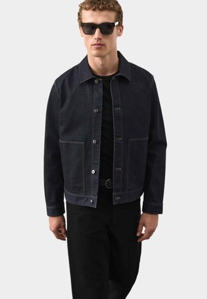 Mann mit schwarzen Sonnenbrillen, dunkler Jeansjacke mit Knöpfen, schwarzem Hemd und schwarzer Hose vor einem schlichten hellen Hintergrund.