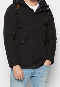 Svart vadderad jacka med huva, med dragkedja och tryckknappsstängning, sidofickor och kontrasterande justeringsdetaljer på huvan.