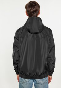 Chaqueta cortavientos negra con capucha, puños elásticos y dobladillo. Hecha de un material liso y ligero, presenta un diseño simple sin logotipos visibles.