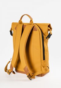 Zaino giallo con chiusura roll-top e dettagli in pelle marrone, spallacci regolabili, tasca laterale con zip e texture del tessuto liscia.