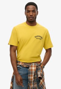 Yellow cotton t-shirt with short sleeves, featuring black "SUPERIOR NEW YORK" text. Worn with light blue jeans and a plaid shirt tied around the waist.