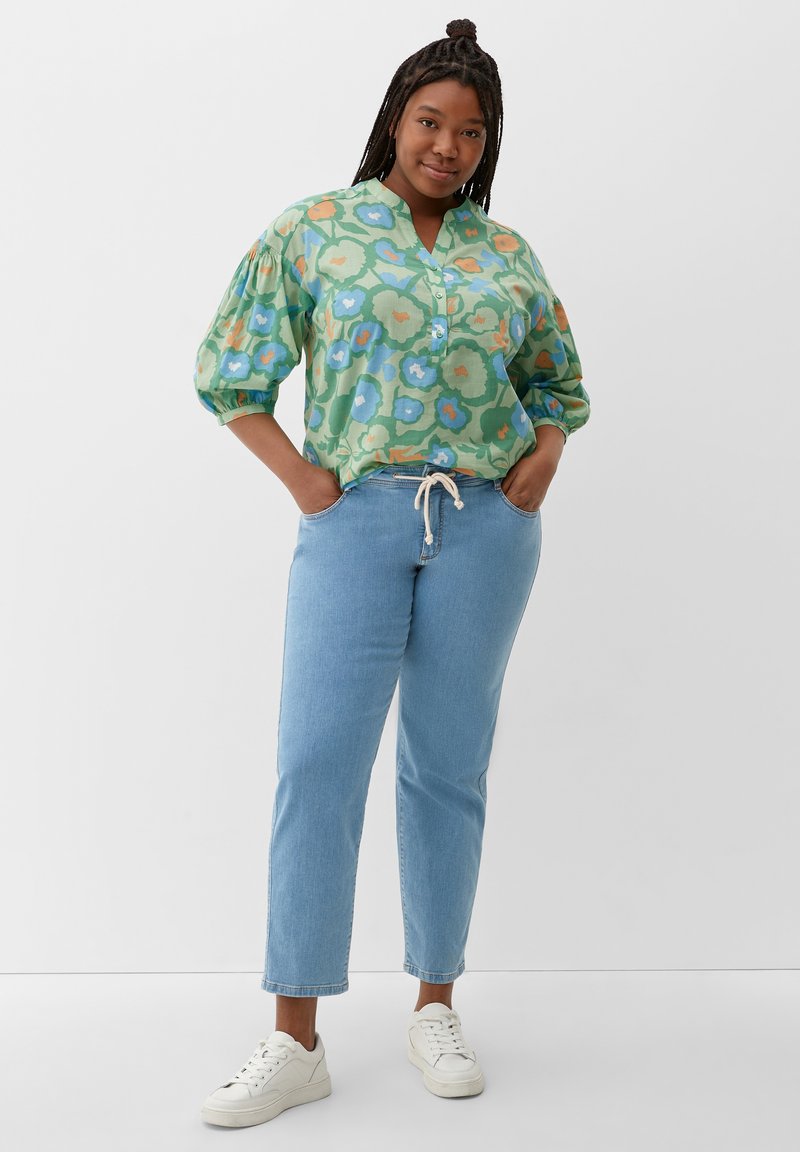 Blusa de estampado floral verde con escote en V y mangas abullonadas, combinada con jeans de mezclilla azul claro y zapatillas blancas. Corte relajado.