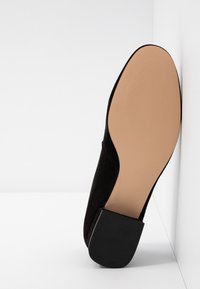 Chaussure en daim noir reposant semelle vers le haut contre un mur blanc, montrant une semelle beige lisse et un talon carré noir.