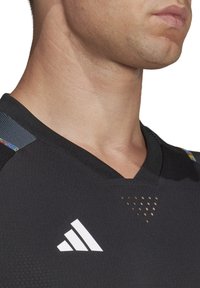 Maglia atletica nera con design a V, dotata di un logo Adidas bianco e accenti colorati sulle spalle. Tessuto testurizzato con fori di ventilazione.