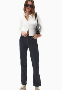 Cárdigan blanco de manga larga con botones en la parte delantera, combinado con jeans rectos de mezclilla oscura y zapatillas beige, usado con gafas de sol negras.