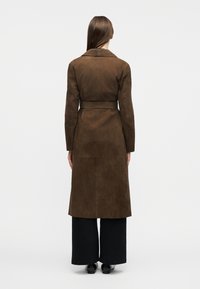 Femme aux longs cheveux bruns portant un manteau en suède marron ceinturé, un pantalon large noir et des chaussures noires, debout de dos sur un fond blanc.