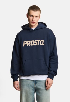 Młody mężczyzna noszący granatową bluzę z kapturem z przednią kieszenią i napisem "PROSTO.", w połączeniu z jasnoniebieskimi dżinsami na jednolitym tle.