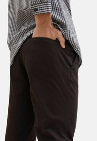 Pantalons en tissu sombre avec poches latérales, associés à une chemise à carreaux. Une main visible dans la poche, soulignant un ajustement décontracté.